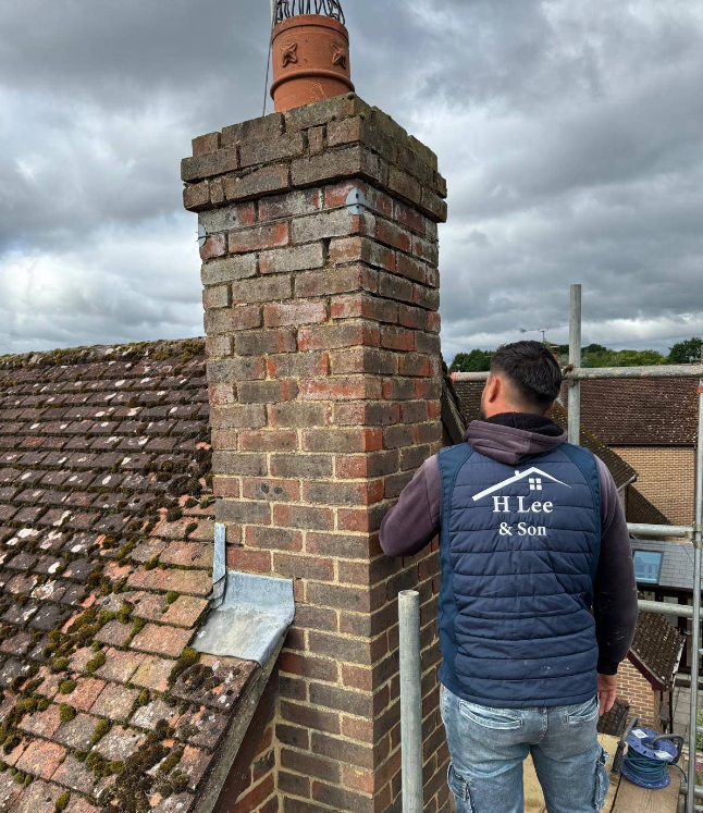 H Lee & Son roofer in branded workwear repointing a brick chimney stack from scaffolding on a tiled roof in Sandhurst